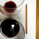 A pair of wine glasses viewed from above. One glass has less red wine than the other. A charcoal pencil rests to the right of the glassware.